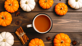 Tasse de café chaud entourée de petites citrouilles orange et blanches, bâtons de cannelle et anis étoilé sur une table en bois, ambiance automnale.