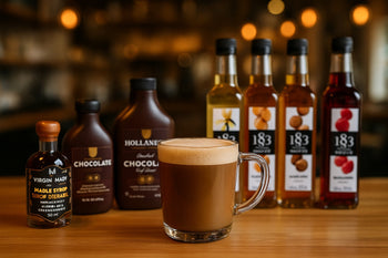 Tasse de café latté dans une tasse en verre, posée sur un comptoir en bois, entourée de sirops 1883, sauces chocolat Hollander et sirop d’érable Virgin Mady, dans une ambiance chaleureuse de café.